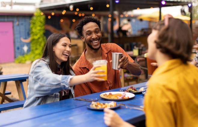 Two women and a man cheering drinks over plates of tacos.