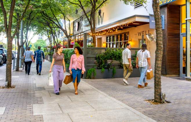 Two women walking down the sidewalk under trees on 2ND Street District with shopping bags