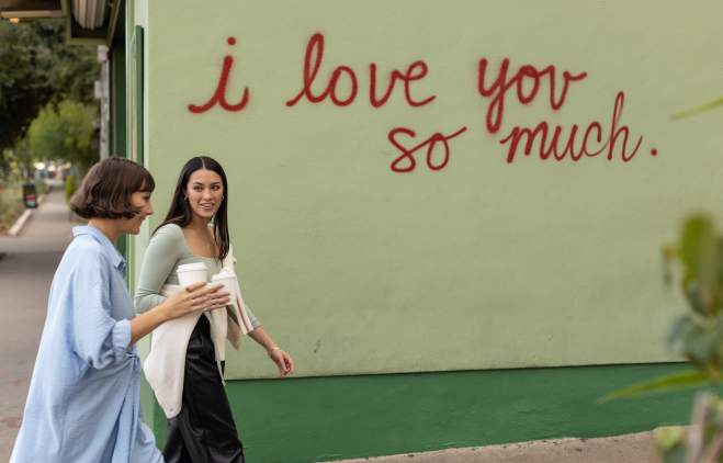 Two women walking in front of the I Love You So Much mural with coffee in hand.