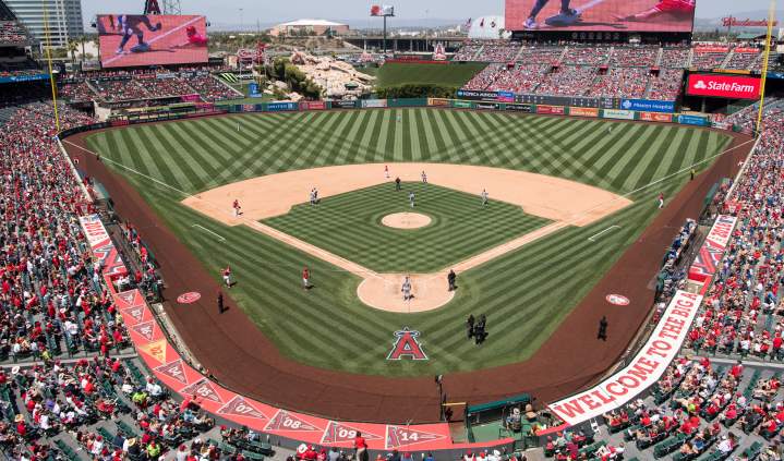 Angel Stadium in Anaheim featuring a wide aerial view of a baseball game in progress, with the diamond field centered, players positioned on the field, large digital screens displaying visuals, and stadium seating filled with spectators on all sides.