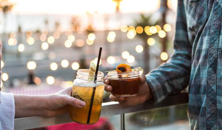 Pacific City in Huntington Beach, California, featuring two people holding handcrafted cocktails on an outdoor patio, with warm string lights and a coastal sunset atmosphere in the background.