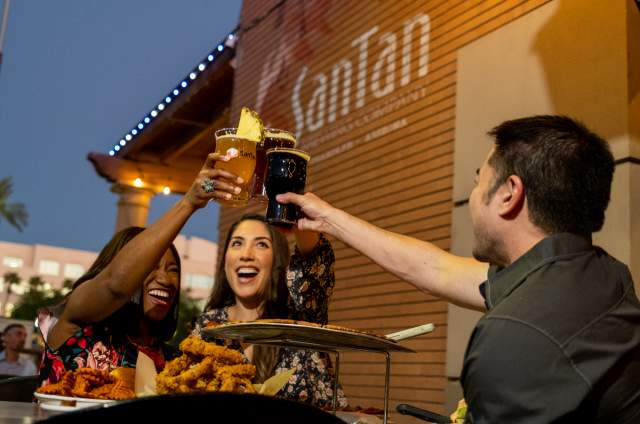 Two women and one man cheers beers on SanTan Brewing patio with food.