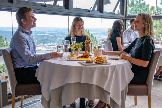 Three diners sit around a table and enjoy a meal at Flagstaff House during the daytime with views of Boulder in the background through the floor-to-ceiling windows