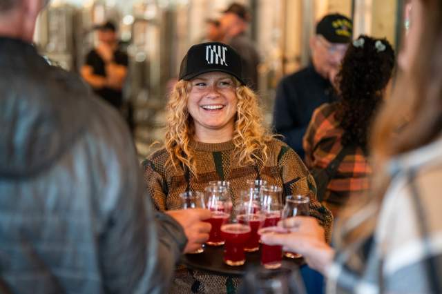 Smiling brewery staff member carrying a tray of red beer samples while guests gather inside a busy taproom at Pattern Break Brewing