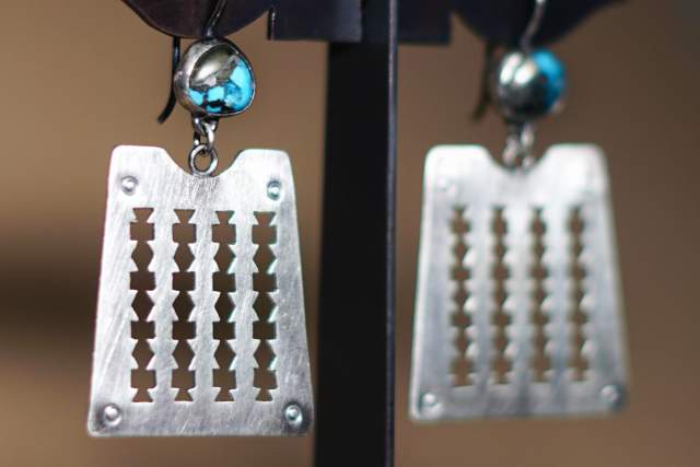 A pair of earrings featuring rectangular pendants with cut-out geometric designs, each adorned with a single turquoise bead, displayed on a black metal stand.