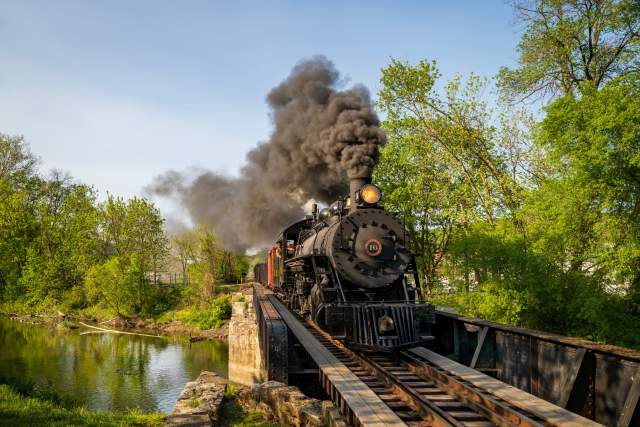 A large, historic train with exhaust coming out the top as it moves across train tracks above a river