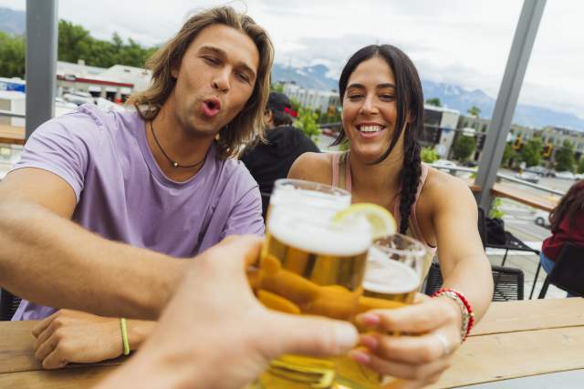 A man and a woman toasting beers with a 3rd person off camera. In the background is Salt Lake City and the wastach mountains