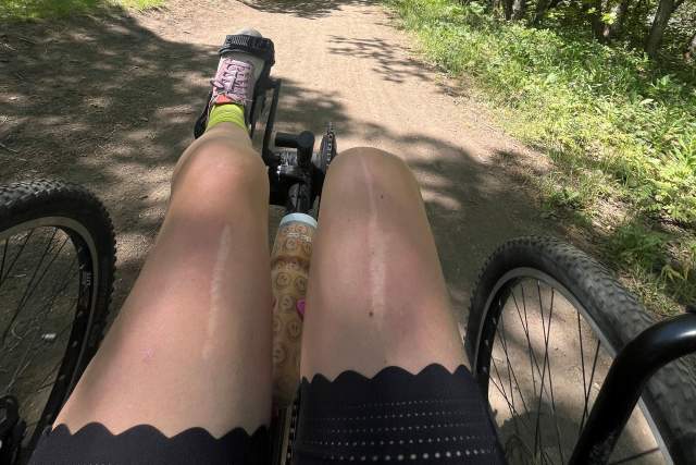 Wide natural trail with trees and grasses on each side. A recumbent quad cycle is biking down the path.