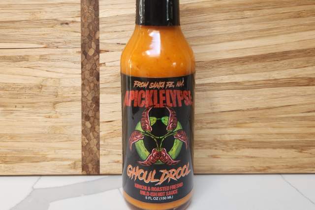 A bottle of Apicklelypse Hot Sauce sits in front of a wooden backdrop