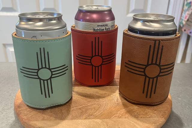 Three cans are placed on a round wooden tray on a countertop. Each can is wrapped in a sleeve with a design resembling the Zia symbol.