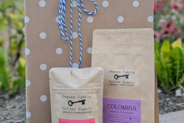 Two bags of coffee labeled "Chavez Family Coffee Supply" from Ethiopia and Colombia, placed next to a polka-dotted brown paper bag tied with blue and white string, outdoors with plants in the background.