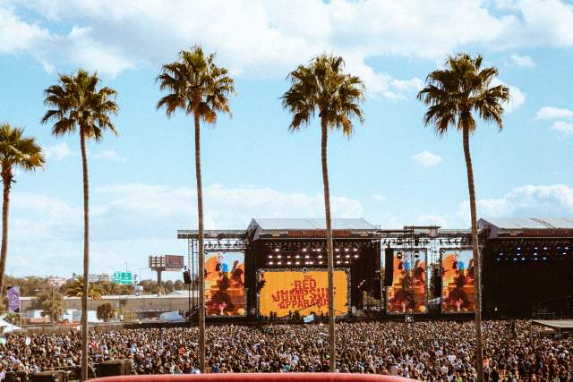 A large crowd gathers at the Vans Warped Tour for an outdoor concert with a main stage featuring video screens and branding signage. Palm trees frame the scene while attendees watch a live performance in an open-air festival setting.