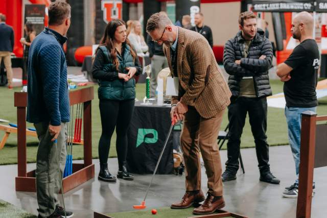 Man putting on a portable mini-golf putting green with people standing around watching