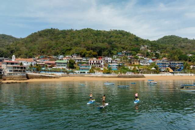 Three people practice paddleboarding in calm waters near the beach and colorful houses on the hillside of Taboga Island, surrounded by lush vegetation.