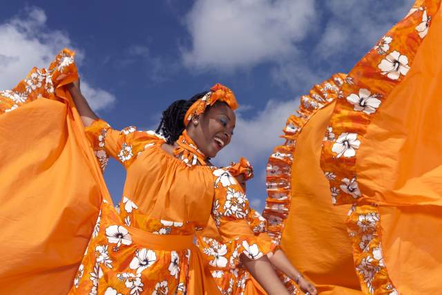 Caribbean dancing troupe wearing bright orange costumes perform on a beach.