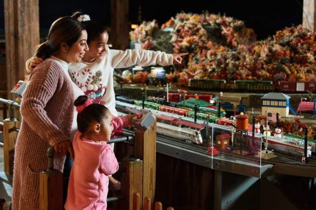 A woman with two small girls observing and pointing at a miniature train display decorated for the holidays at The Brandywine Museum of Art