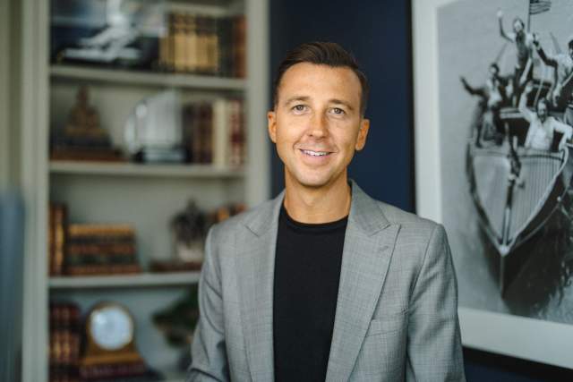 Smiling man in a medium grey suit with a black t-shirt standing in front of a bookshelf and painting