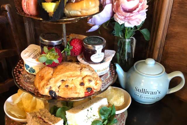 A platter of sandwiches and cakes with a teapot in the background.