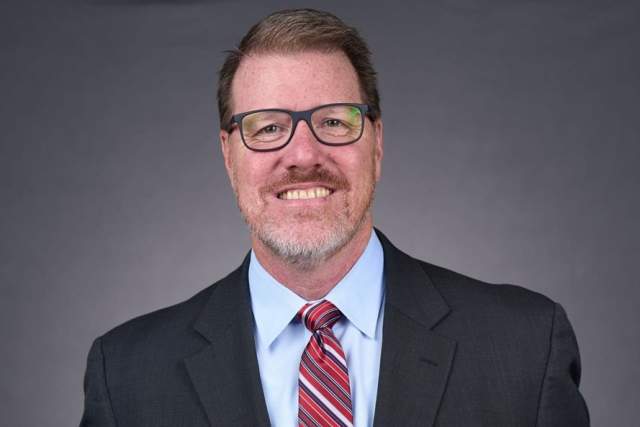 Photo of Scott Draper standing in a dark suit and light blue shirt with a red striped tie