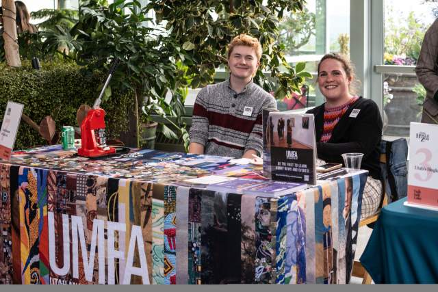 Vendor table covered in a colorful cloth that says UMFA, has swag on it and 2 smiling people sitting behind it.