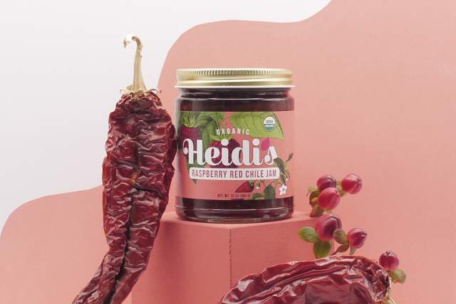A red jar sits atop a red block with red chile and raspberries surrounding the base. A red design sits behind it on a white wall. The label reads "Organic, Heidi's Red Raspberry Chile Jam