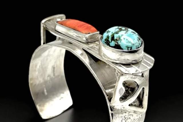 Silver cuff bracelet featuring a turquoise stone and an orange stone, with geometric cut-out designs.