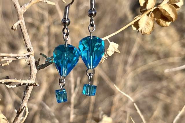 A pair of turquoise crystal earrings hanging from a branch with dried leaves in a natural setting.