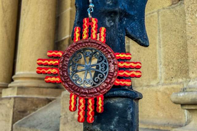 A decorative ornament featuring a red and yellow Zia symbol with a star shape at the center hangs on a black wrought iron fixture. The background shows the textured stone of a building. A "NM True Certified" badge appears in the corner.