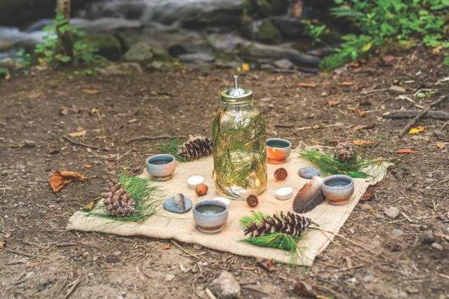 A serene outdoor tea setup beside a small forest waterfall. A burlap cloth is laid on the forest floor, topped with a glass jar filled with pine needles and water, surrounded by ceramic cups, pinecones, pebbles, and sprigs of evergreen. The background shows lush green foliage and a softly flowing creek cascading over rocks.