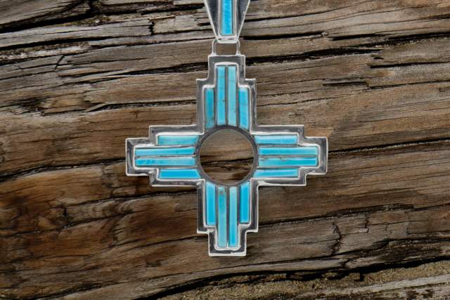 Turquoise inlaid zia pendant with a circular center and symmetrical design, on a wooden background.