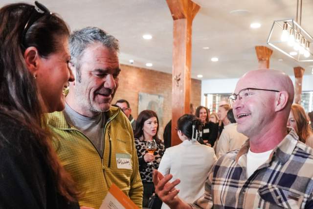 Three people standing, smiling and talking to each other. In the background are more groups of people conversing