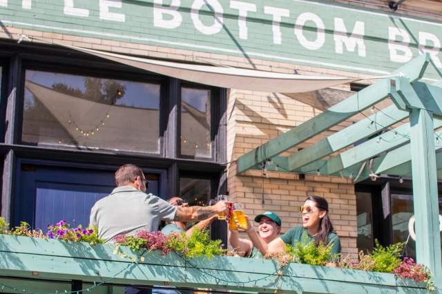 People on a patio of a restaurant cheersing glasses of beer under a sign that reads Triple Bottom Brewery