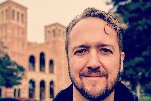 Sepia toned image of man in leather jacket with historic looking buildings in the background