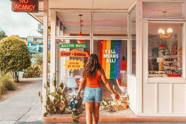 Girl walking away from the camera in front of the window at Austin Motel with a Pride flag hanging in the window with the words "Y'all Mean All".