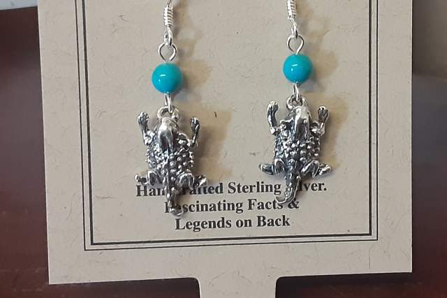 Sterling silver earrings featuring frog designs and blue beads, displayed on a card labeled "The Horny Toad Collection" which mentions handcrafted details and additional information on the back.