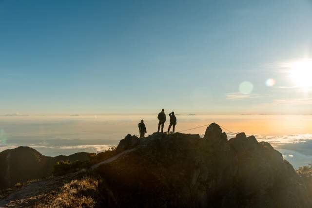The image captures the silhouette of three people on the summit of a rugged mountain, likely Volcán Barú, at dawn or dusk, with the sun shining intensely to one side. Below them, a sea of clouds stretches to the horizon, creating a breathtaking landscape and an atmosphere of majestic tranquility.