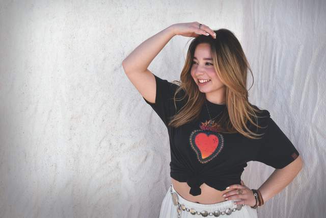 A woman wearing a black shirt with a red heart standing against a white textured wall