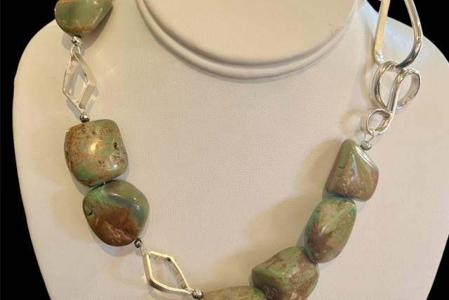 A necklace featuring polished green and turquoise gemstones and silver links displayed on a white mannequin bust.