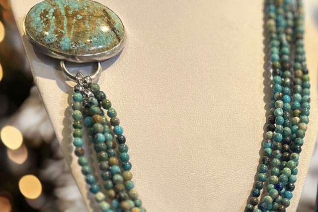 An elegant multi-strand turquoise bead necklace with a large oval pendant, displayed on a white bust form against a blurred background of warm lights and pine branches.