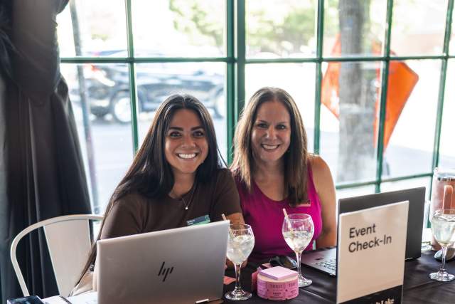 Two smiling women sitting at a table that has a runner on it that says Visit Salt Lake. They have computers set up for registration.