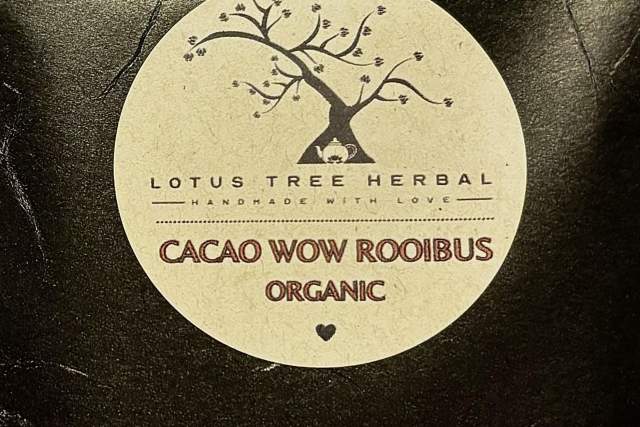 A package of Lotus Tree Herbal Cacao Wow Rooibos Organic tea, resting against a decorative twisted wire tree sculpture, set against a textured red background.