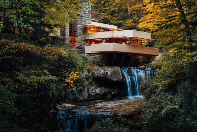 A modern, tan home , known as Frank Lloyd Wright's Fallingwater, situated on top of a waterfall with fall foliage-filled trees surrounding it