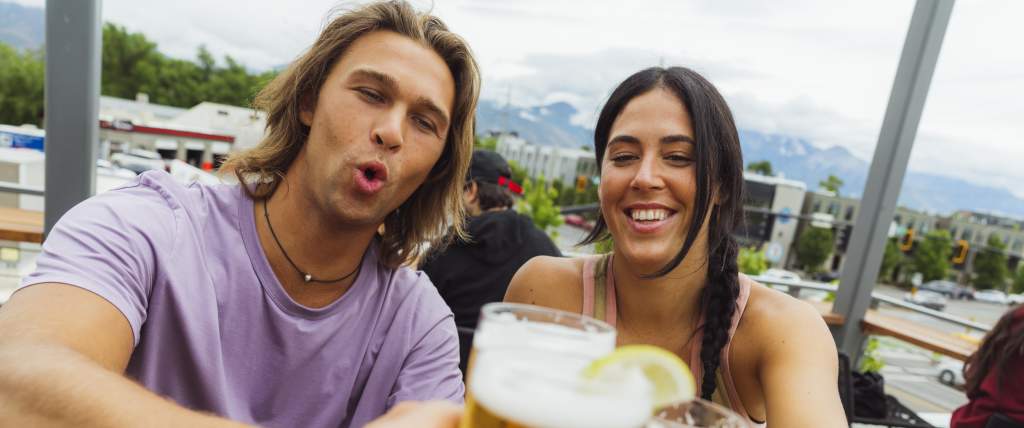 A man and a woman toasting beers with a 3rd person off camera. In the background is Salt Lake City and the wastach mountains