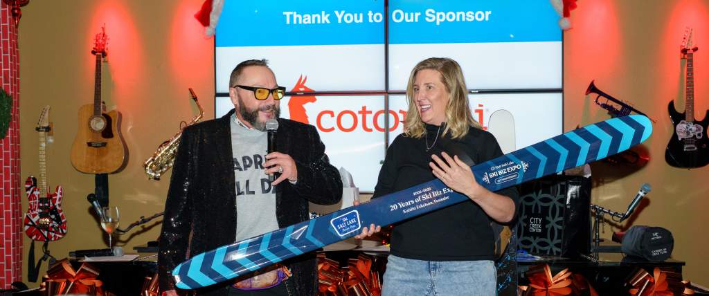 Two people are standing in front of a crowd. One holds a microphone the other is holding dynastar skis