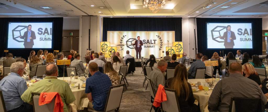 Man in brown suit speaking on a stage with the logo of the SALT Summit behind him and round tables with people attentively listening