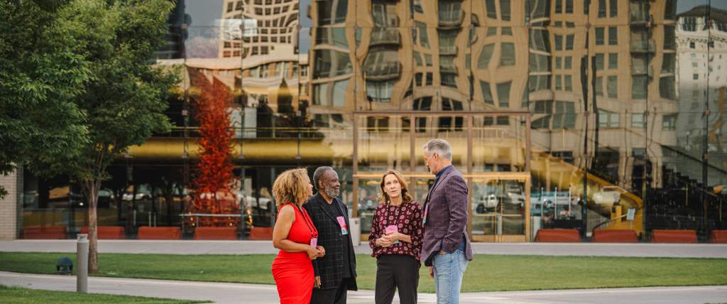 4 Conference attendees with conference badges standing in front of a building with glass reflecting the Salt Lake skyline