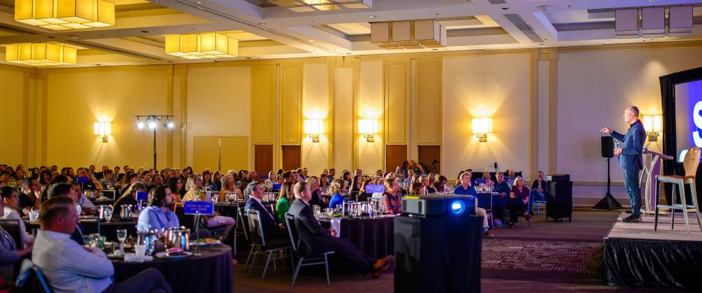 Will Guiderra, a white man in a suit, standing on stage talking to a room full of people sitting at round tables