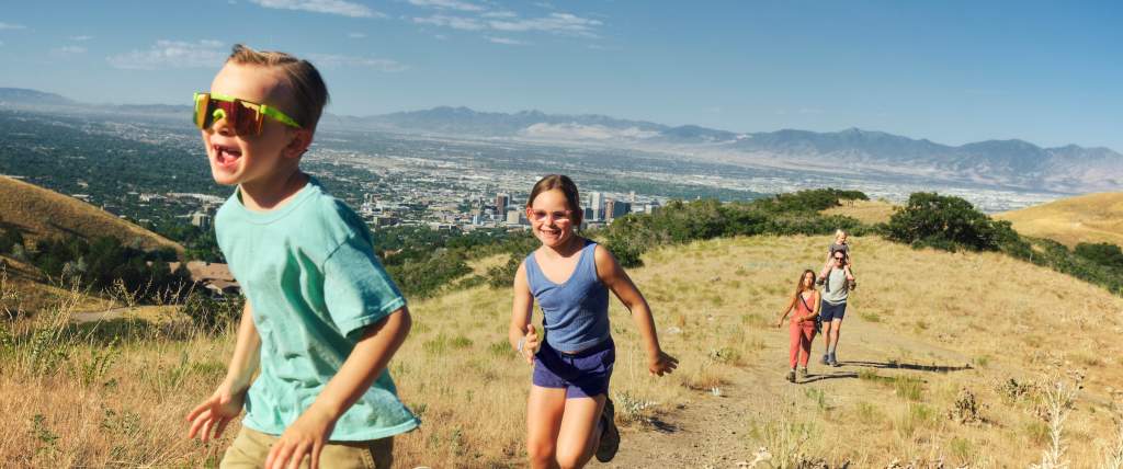 Kids running up a trail on the top of a hill with the Salt Lake valley and Oquirrh mountains in the background