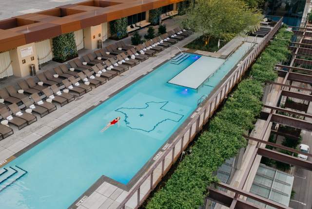 Aerial photo of the JW Marriott rooftop pool with a woman in a red bathing suit swimming on her back in the center of the pool. The floor of the pool includes an outline of the state of Texas with a star on the location of Austin.