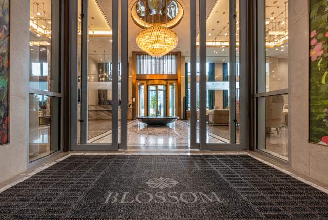 Grand lobby entrance at Blossom Hotel Houston, featuring glass doors opening to a spacious interior with a large golden chandelier, polished marble floors, modern seating areas, and a round decorative water feature centered beneath warm ambient lighting.